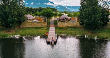 A wooden dock leading to a house with a dome-like structure, surrounded by greenery and a body of water.