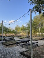 Un área de estar al aire libre con mesas y sillas, rodeada de árboles y un cielo despejado.