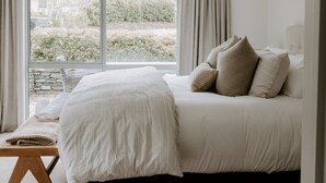 Ein Schlafzimmer mit großem Fenster, einem Bett mit vielen Kissen, einem Nachttisch und einem gewebten Korb.