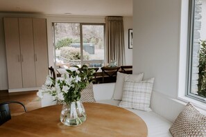 Un coin repas avec une table ronde, des chaises, un vase de fleurs, une fenêtre avec des rideaux et une vue sur l’extérieur.