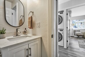 A modern bathroom with a large mirror, a sink, and a toilet. There are two towels hanging on hooks, and a potted plant on the counter. - Park Place (Colorado Springs)
