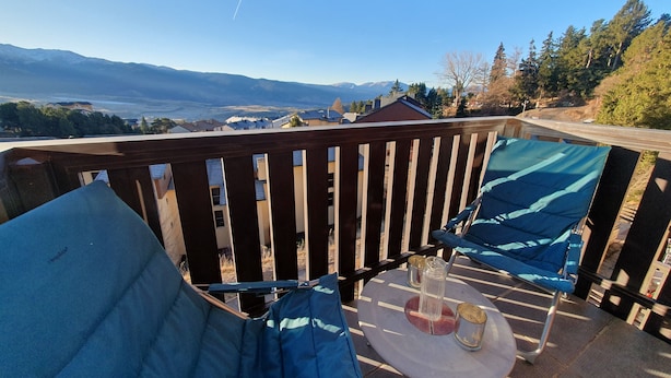 A balcony with a view of mountains and houses, a table with a glass, and a blue chair. - Studio Font-romeu Vue Imprenable Plein sud sur le Cambre D'aze ! (Font-Romeu-Odeillo-Via)