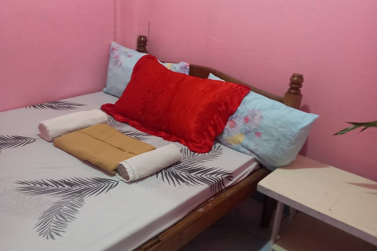 Un lit simple avec une couvre-lit à motifs, un oreiller rouge et une serviette beige.