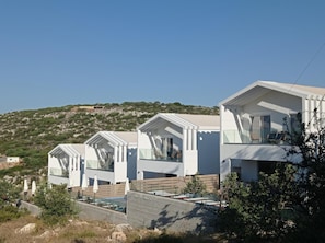 A row of modern, white villas with large windows and balconies, set against a hilly backdrop. - Pearl Nevelo Quality Villas (Rethymno)