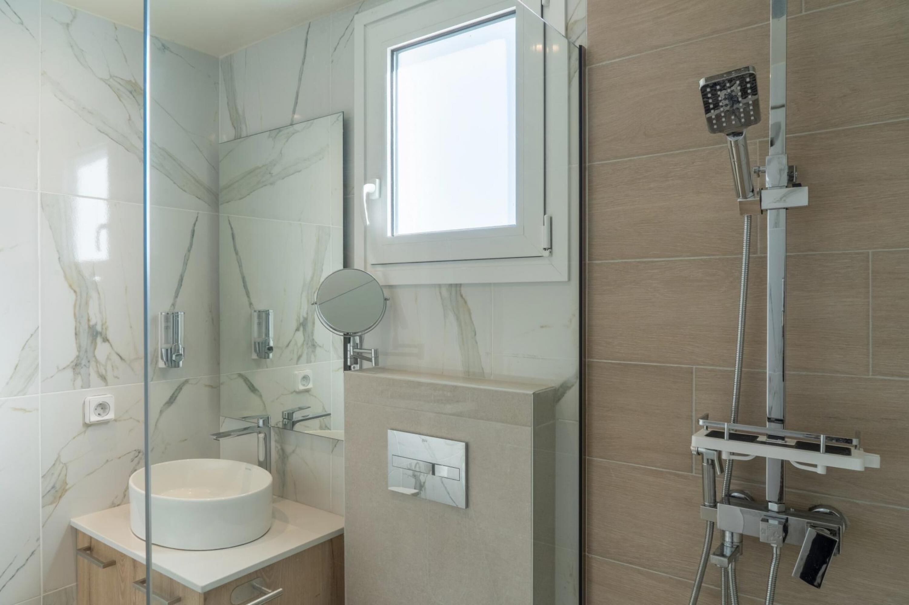 A modern bathroom with a marble vanity, a round mirror, a built-in shower, and a window.