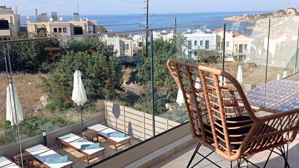 Un balcon agrémenté de fauteuils et d’une table en osier, offrant une vue sur une ville côtière avec ses bâtiments et la mer.