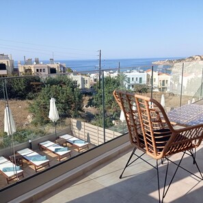A balcony with wicker chairs and a table, overlooking a coastal town with buildings and the sea. - Amethyst Nevelo Quality Villas (Rethymno)