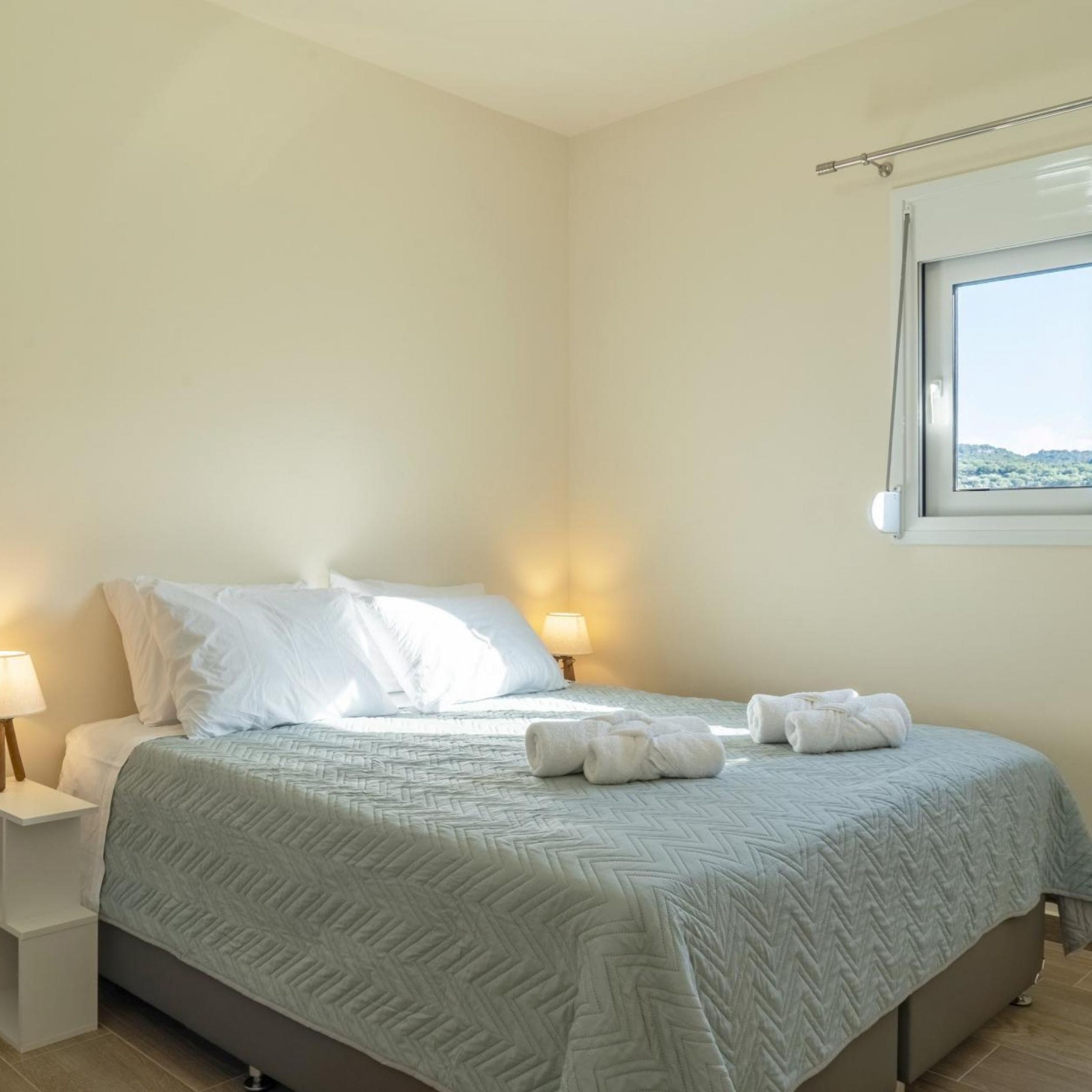 A neatly made bed with a patterned bedspread, two bedside lamps, and a window with a view of greenery.