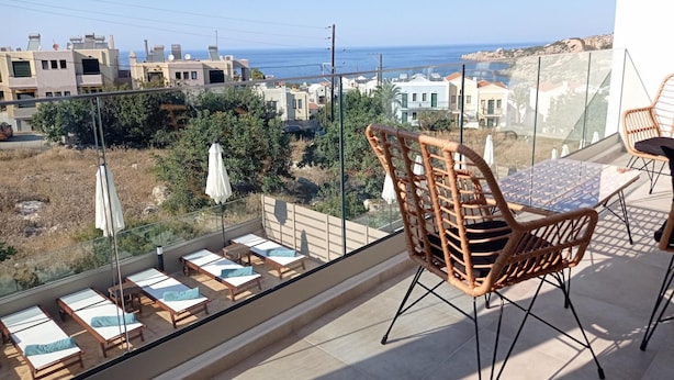 A balcony with sunbeds and a wicker chair, overlooking a coastal town with buildings and the sea. - Diamond Nevelo Quality Villas (Rethymno)