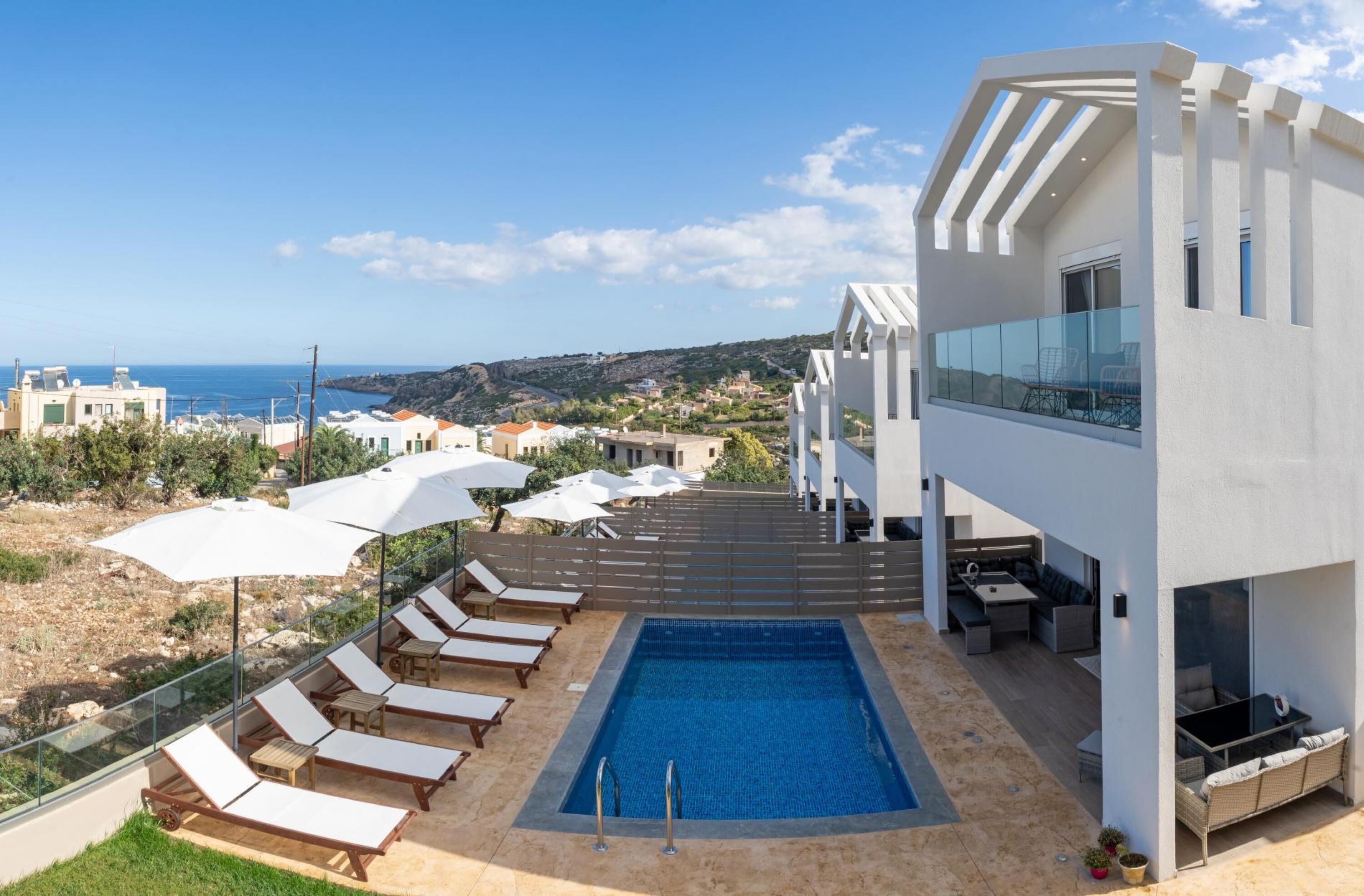 A modern outdoor pool area with lounge chairs and umbrellas, a clear view of the sea, and a contemporary building with a balcony.
