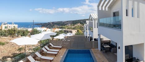 A modern outdoor pool area with lounge chairs and umbrellas, a clear view of the sea, and a contemporary building with a balcony.