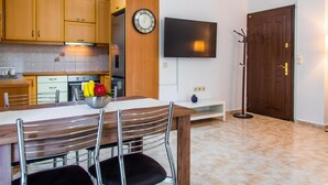 A modern kitchen with wooden cabinets, a dining table with chairs, a fruit bowl, a wall-mounted clock, a flat-screen TV, and a coat rack. - Kantouni 2 Heraklion Apartment (Heraklion)