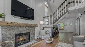 Un vaste salon doté d’une cheminée en pierre, d’un grand téléviseur à écran plat, d’une table basse en bois et d’un escalier menant à un niveau supérieur.