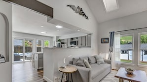 Un salon moderne avec un canapé gris, une petite table d’appoint en bois et une vue sur l’extérieur grâce à de grandes fenêtres.
