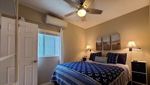 A bedroom with a bed, a ceiling fan, a window with blinds, a nightstand, a lamp, and artwork on the wall. - Waypoint Cottage, one block from the beach! (San Diego)