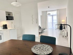 A modern kitchen with white cabinetry, a microwave, and a coffee machine. There is a dining area with a wooden table and teal chairs. - Wit II, Near Metro, Train N7670 (Brussels)