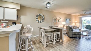 A kitchen with white cabinets, a dining area with a wooden table and white chairs, a striped armchair, and a sofa. - Pawleys Island Condo w/ Screened Porch + Golfing! (Pawleys Island)