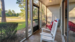 A covered porch with a view of a golf course, a white bench, and a red chair. - Pawleys Island Condo w/ Screened Porch + Golfing! (Pawleys Island)