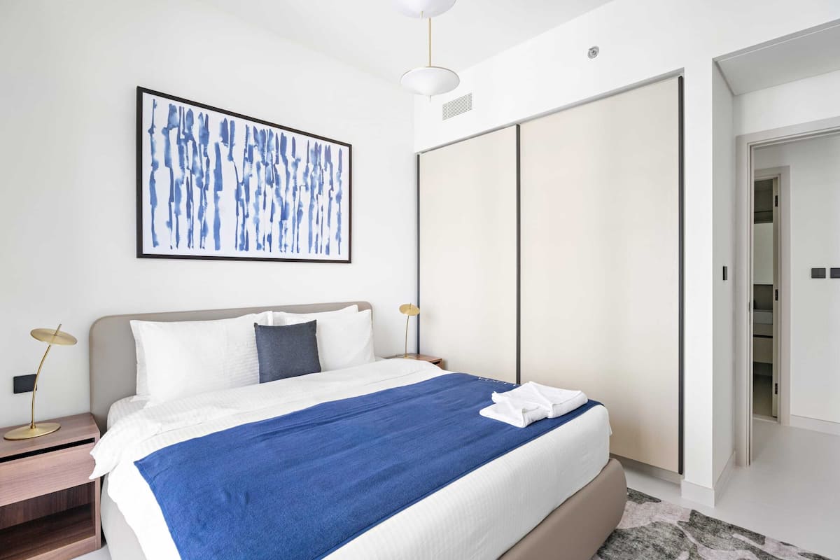 Una habitación de hotel moderna con una cama grande, una mesita de noche con lámpara, un armario y un cuadro abstracto enmarcado en la pared.