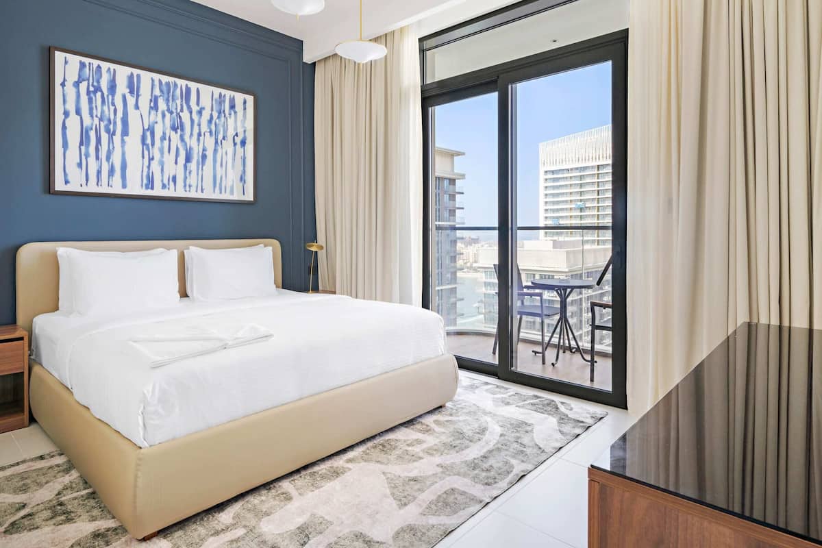 Una habitación de hotel moderna con una cama grande, vistas de la ciudad a través de ventanales del suelo al techo y un balcón con mobiliario exterior.