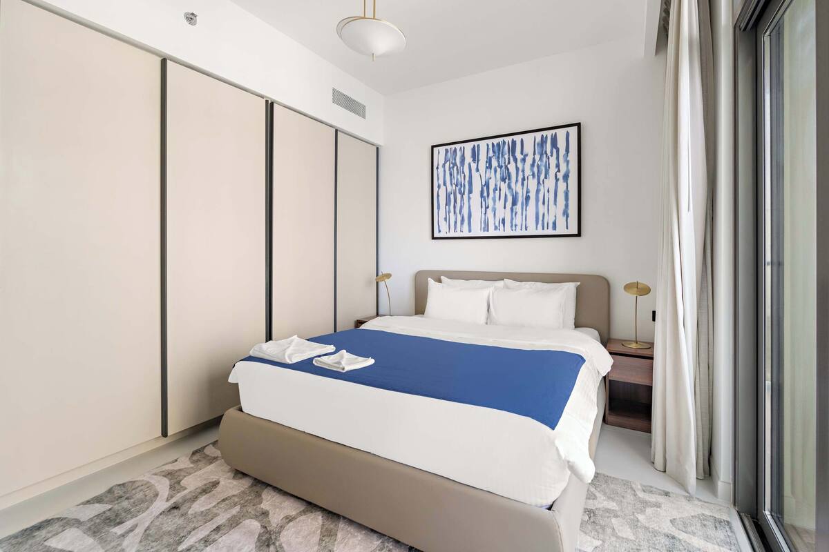 Una habitación de hotel moderna con una cama grande, un armario, una mesita de noche con lámpara y un ventanal con cortinas.