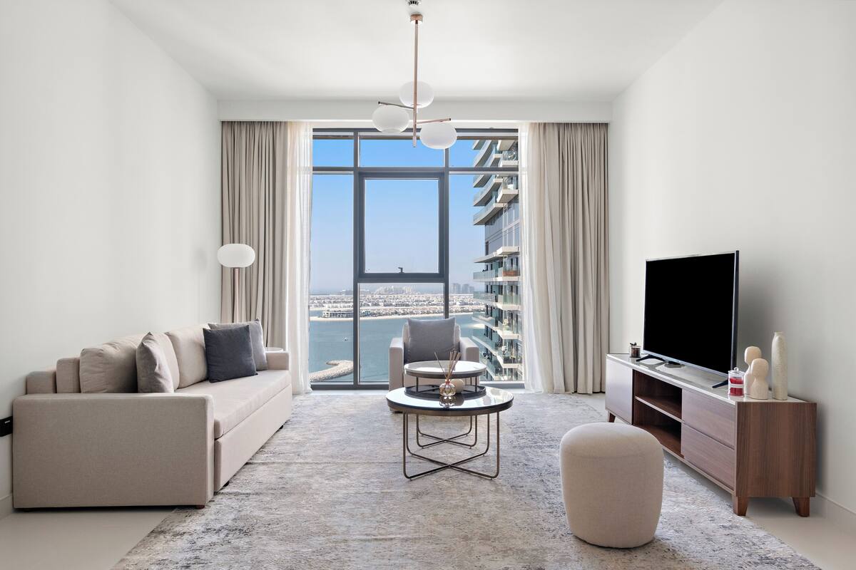 Una sala de estar moderna con un sofá, una mesa de centro, un televisor y vistas a un paisaje urbano a través de grandes ventanales.