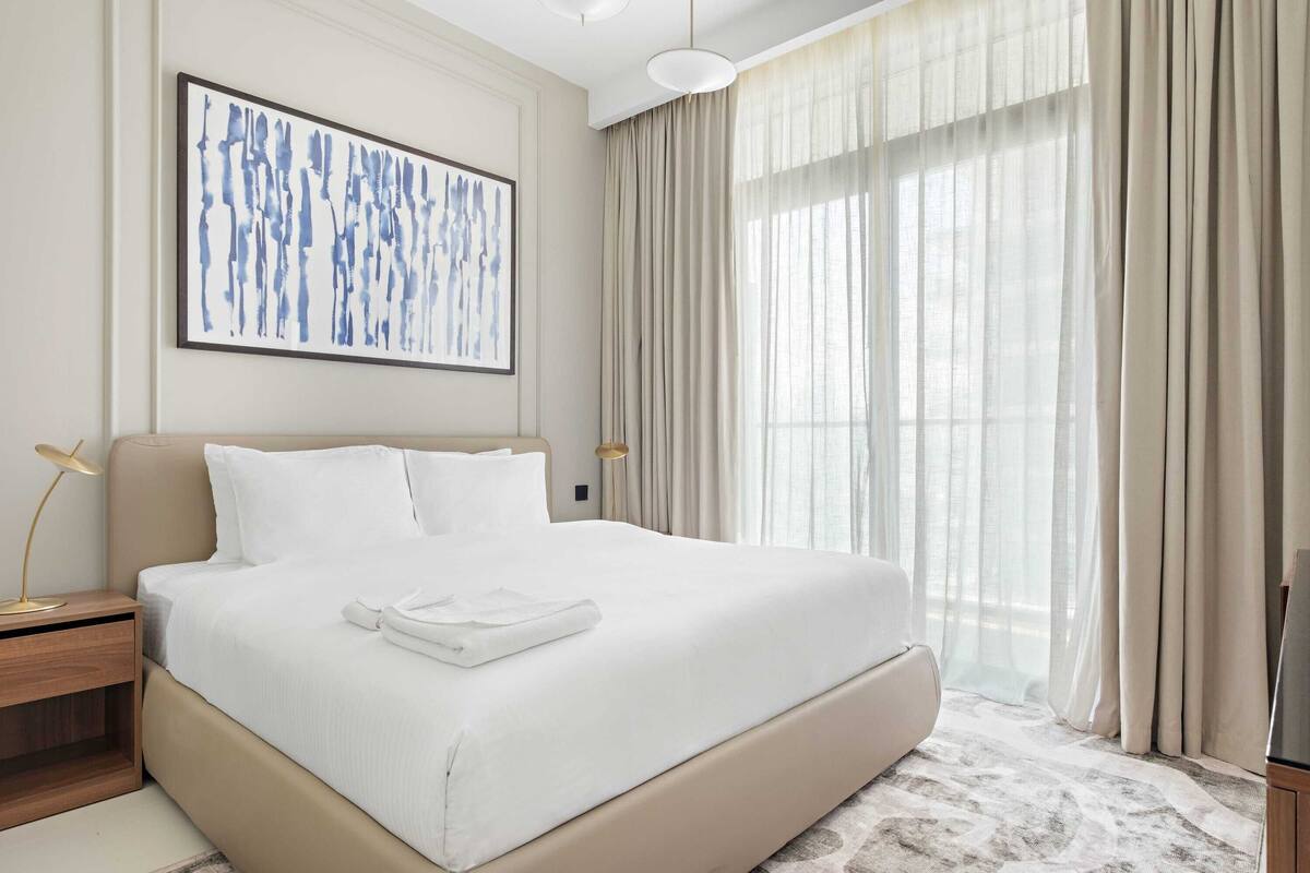 Una habitación de hotel con una cama grande, un cuadro abstracto enmarcado, una lámpara de noche y cortinas translúcidas.