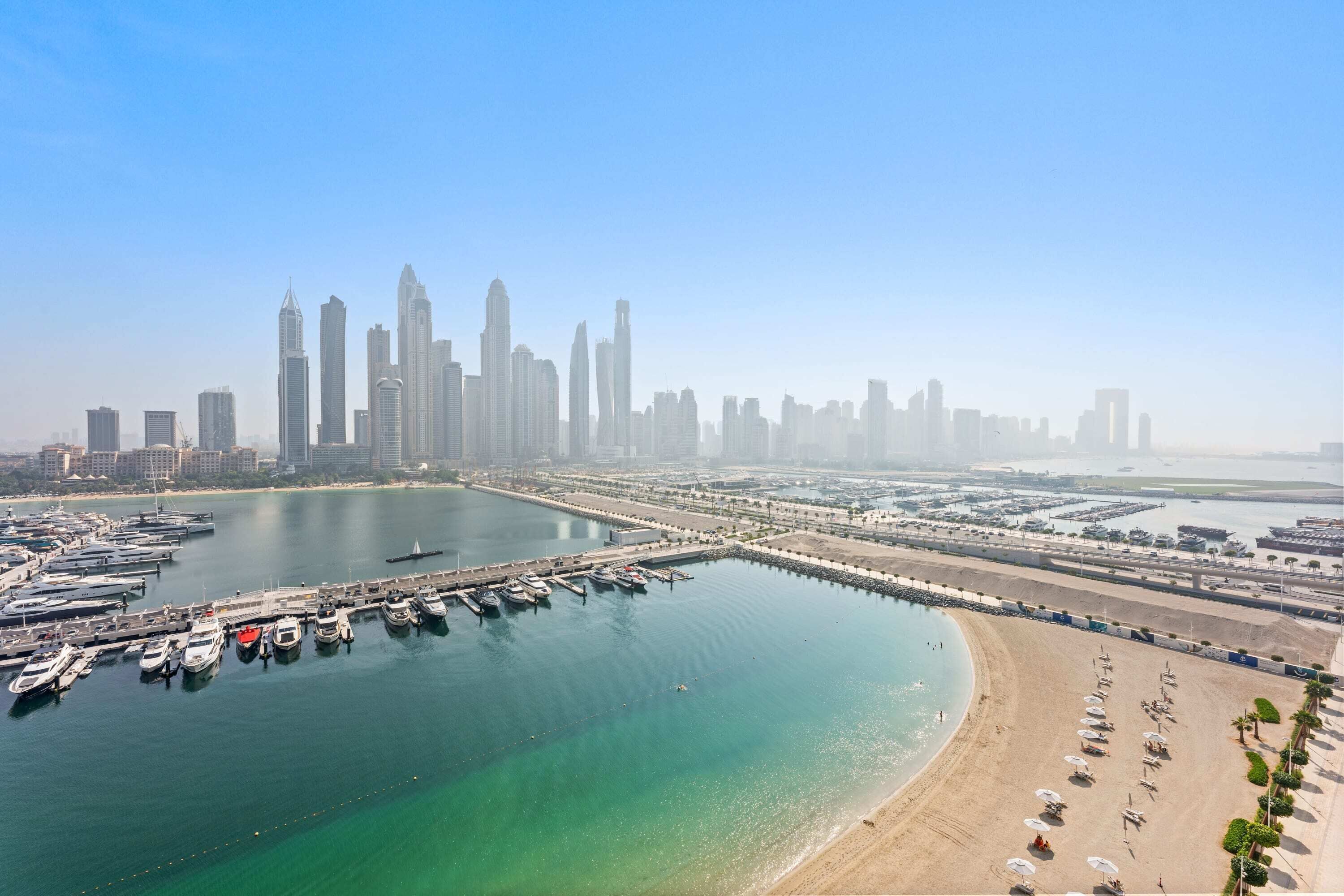 A marina with boats docked, a beach with umbrellas, and a city skyline in the background.
