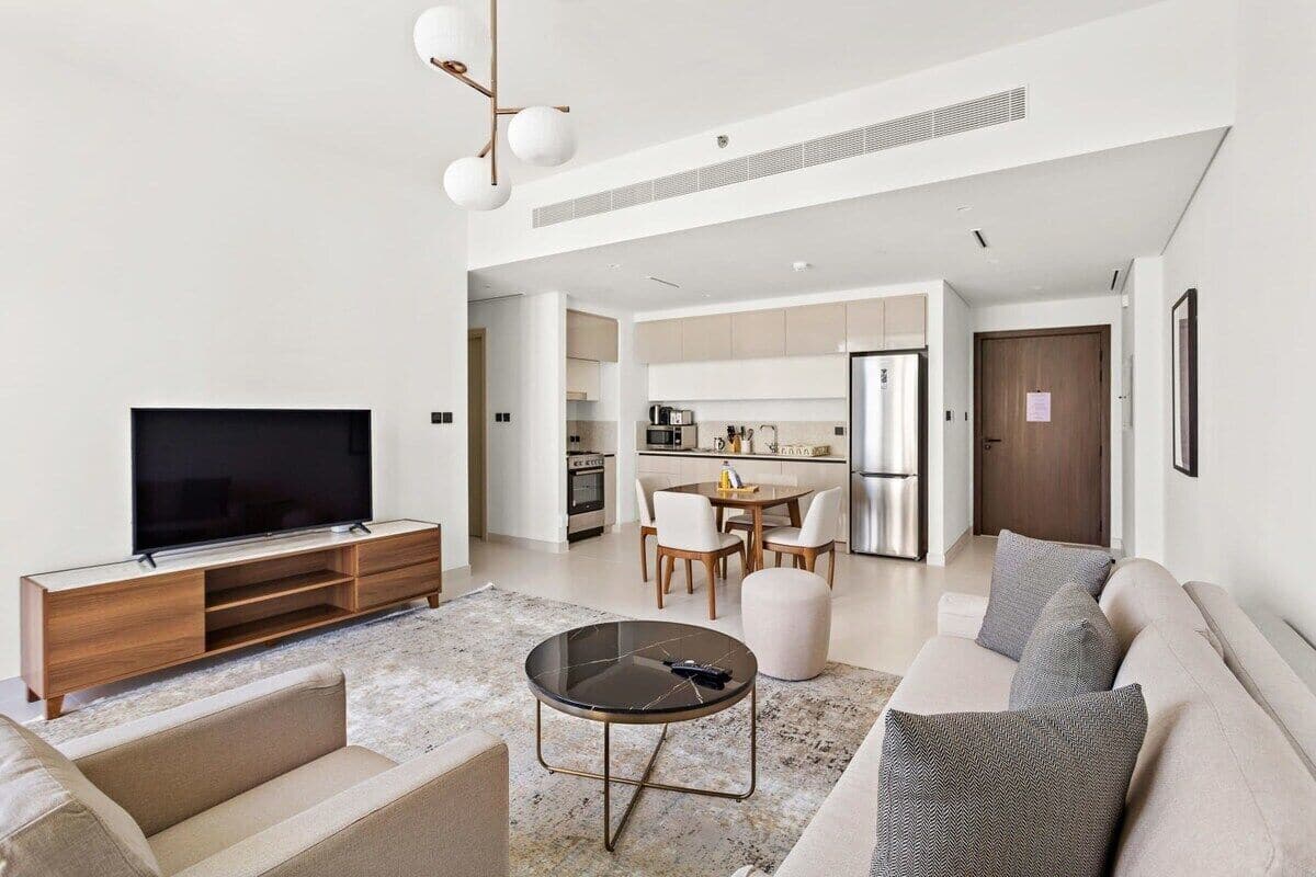 Una sala de estar moderna con una televisión de pantalla plana, una mesa de centro redonda y un sofá. Se ve un área de cocina con una mesa de comedor y sillas al fondo.