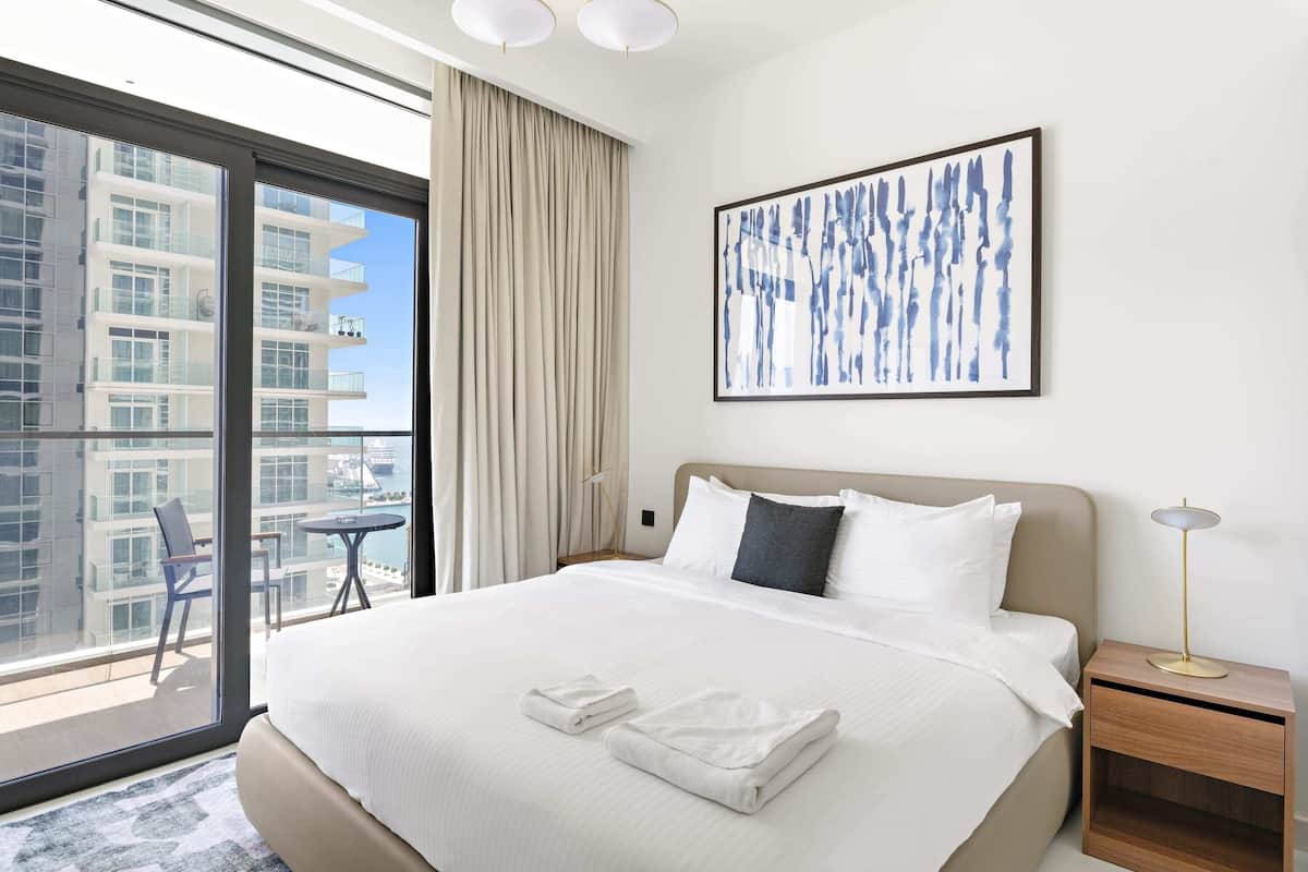 Una habitación de hotel con una cama grande, vistas de la ciudad, una obra de arte moderno y una mesita de noche con lámpara.