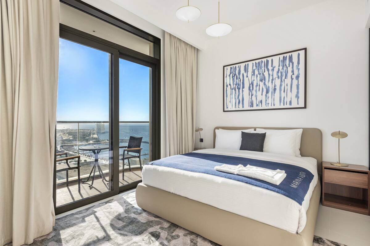 Habitación de hotel con una cama grande, vistas de la ciudad a través de ventanales del suelo al techo, un balcón con mesa y sillas, y un cuadro abstracto enmarcado en la pared.