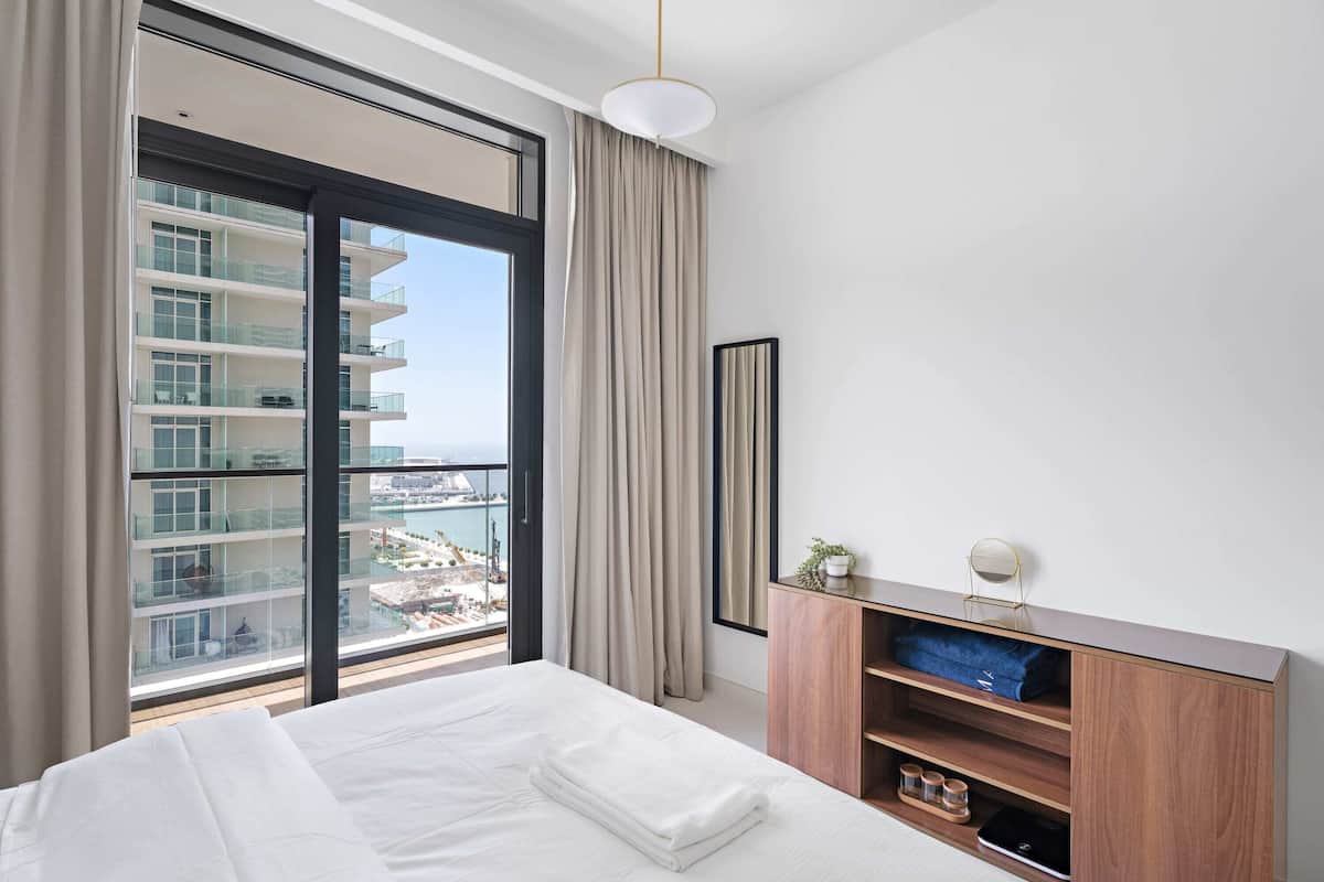 Una habitación de hotel moderna con una cama grande, una mesita de noche de madera, vistas de la ciudad a través de ventanales del suelo al techo, y un balcón con vista al puerto deportivo.