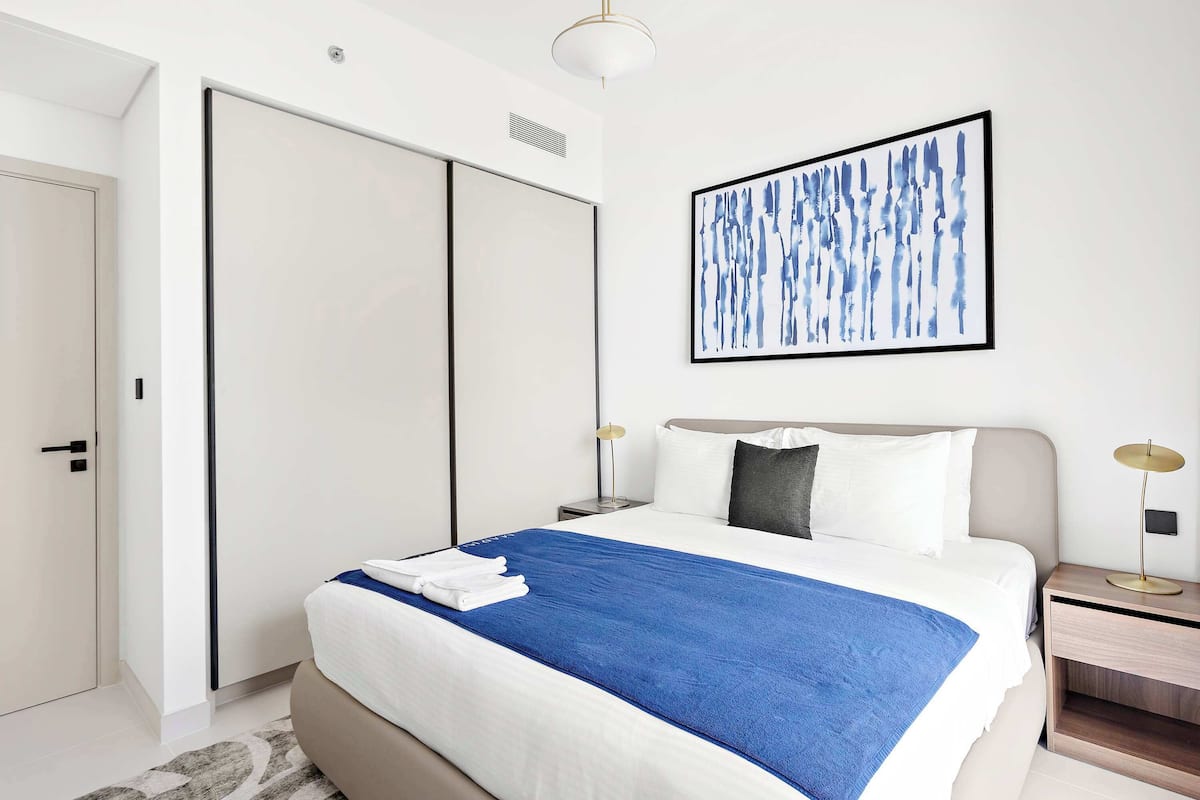 Una habitación de hotel moderna con una cama grande, un armario, una mesita de noche con lámpara y un cuadro abstracto en la pared.
