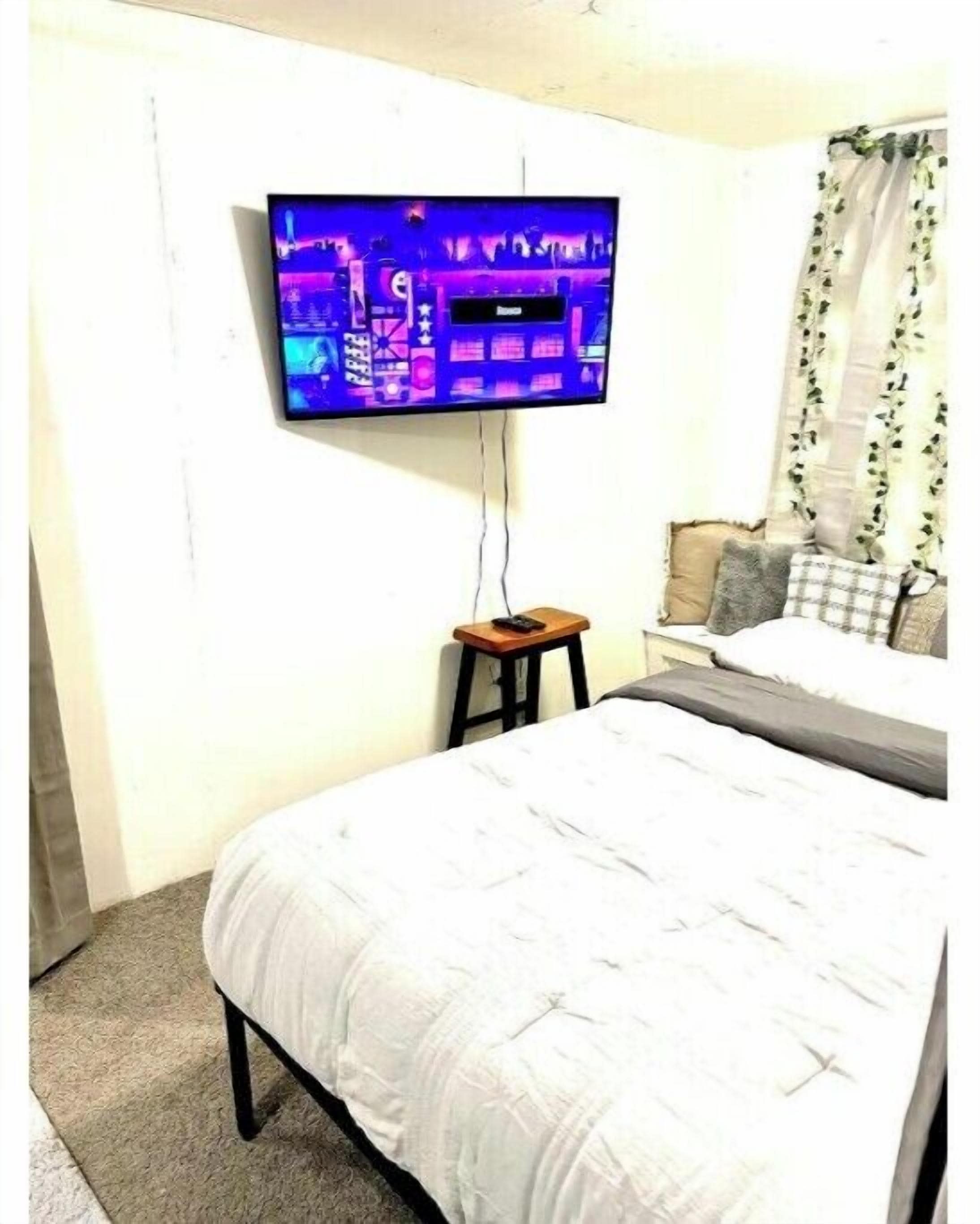 Een slaapkamer met een bed, een aan de muur bevestigde televisie, een klein houten krukje en een raam met gordijnen.