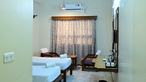Habitación de hotel con dos camas individuales, una mesita pequeña, una silla, un escritorio, un equipo de aire acondicionado, un ventilador de techo y una ventana con cortinas.