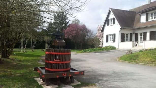 A rural road with a large cylindrical structure on a trailer, surrounded by greenery and a white house with dark shutters. - La Villa Champenoise (Pierry)