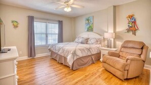 A bedroom with a bed, a chair, a ceiling fan, a window with curtains, and wall art. - 0.3 mi. to beach, 4 BR, 3 Bath, sleeps 9, grill, pool table, games, LSV for rent (North Myrtle Beach)