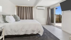 A bedroom with a bed, a nightstand, a window with curtains, an air conditioning unit, and a sliding glass door leading to a balcony. - Sutherland Sunrise - Kingscliff Studio (Tweed)