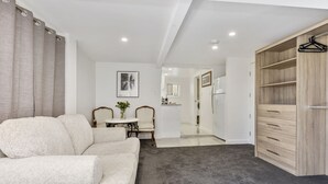 A modern living room with a sofa, a small table with chairs, a wooden wardrobe, and a kitchen area with a refrigerator. - Sutherland Sunrise - Kingscliff Studio (Tweed)