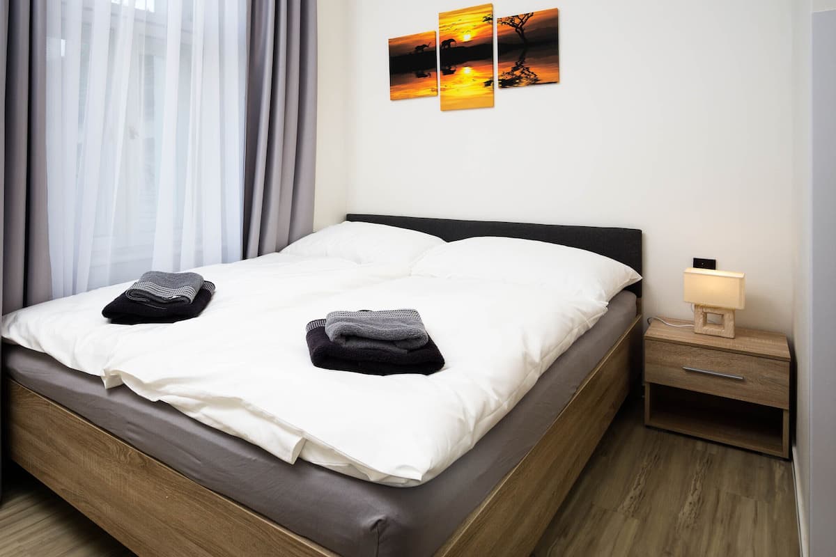 A neatly made bed with two folded towels, a bedside table with a lamp, and two wall art pieces depicting a sunset.