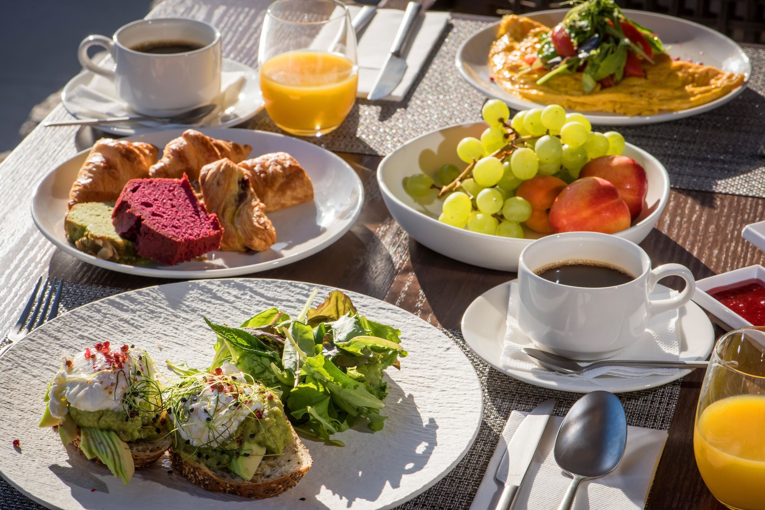A breakfast table with croissants, a bowl of grapes and tomatoes, and a plate of eggs with greens.