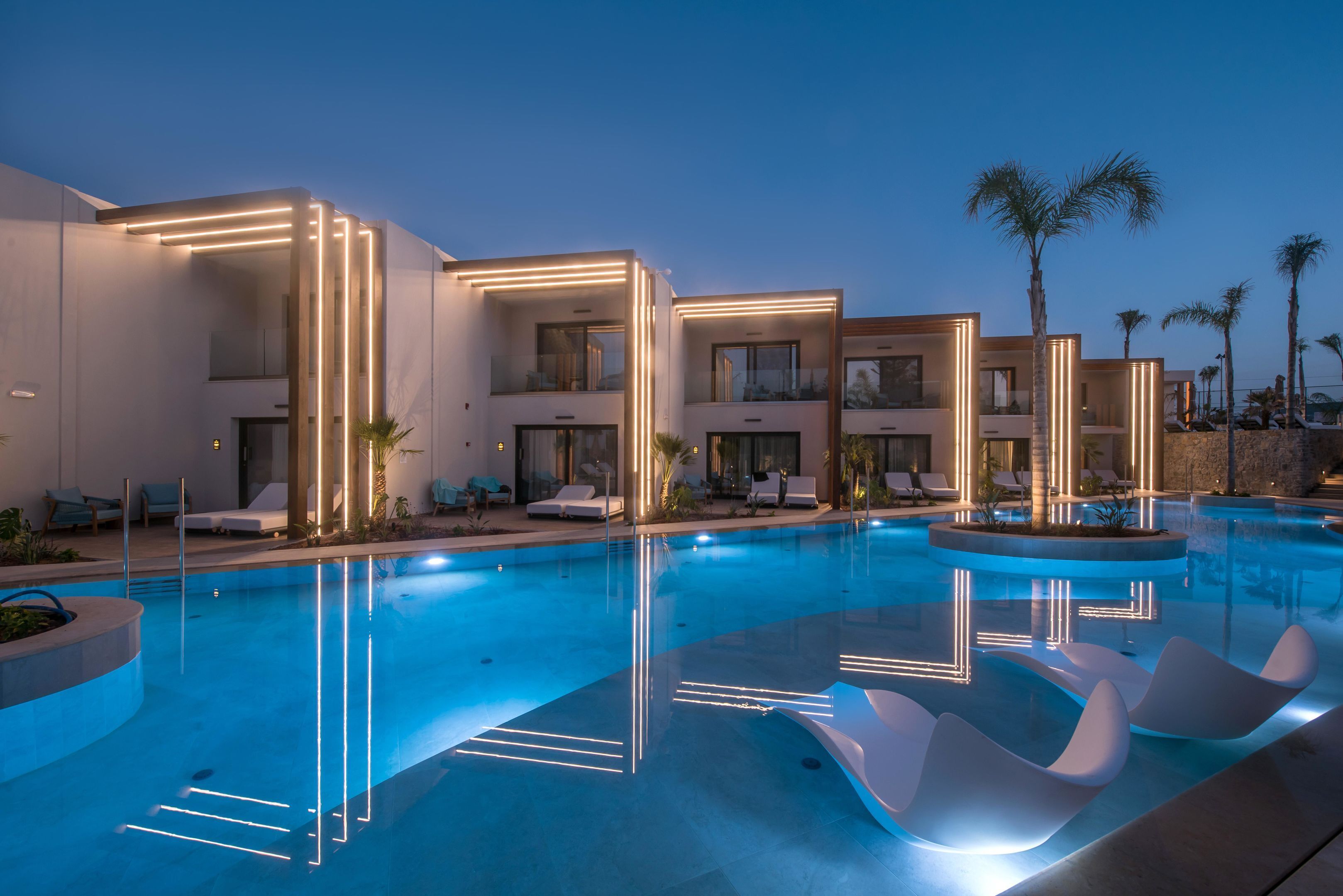 A modern hotel with a swimming pool, lounge chairs, and palm trees.