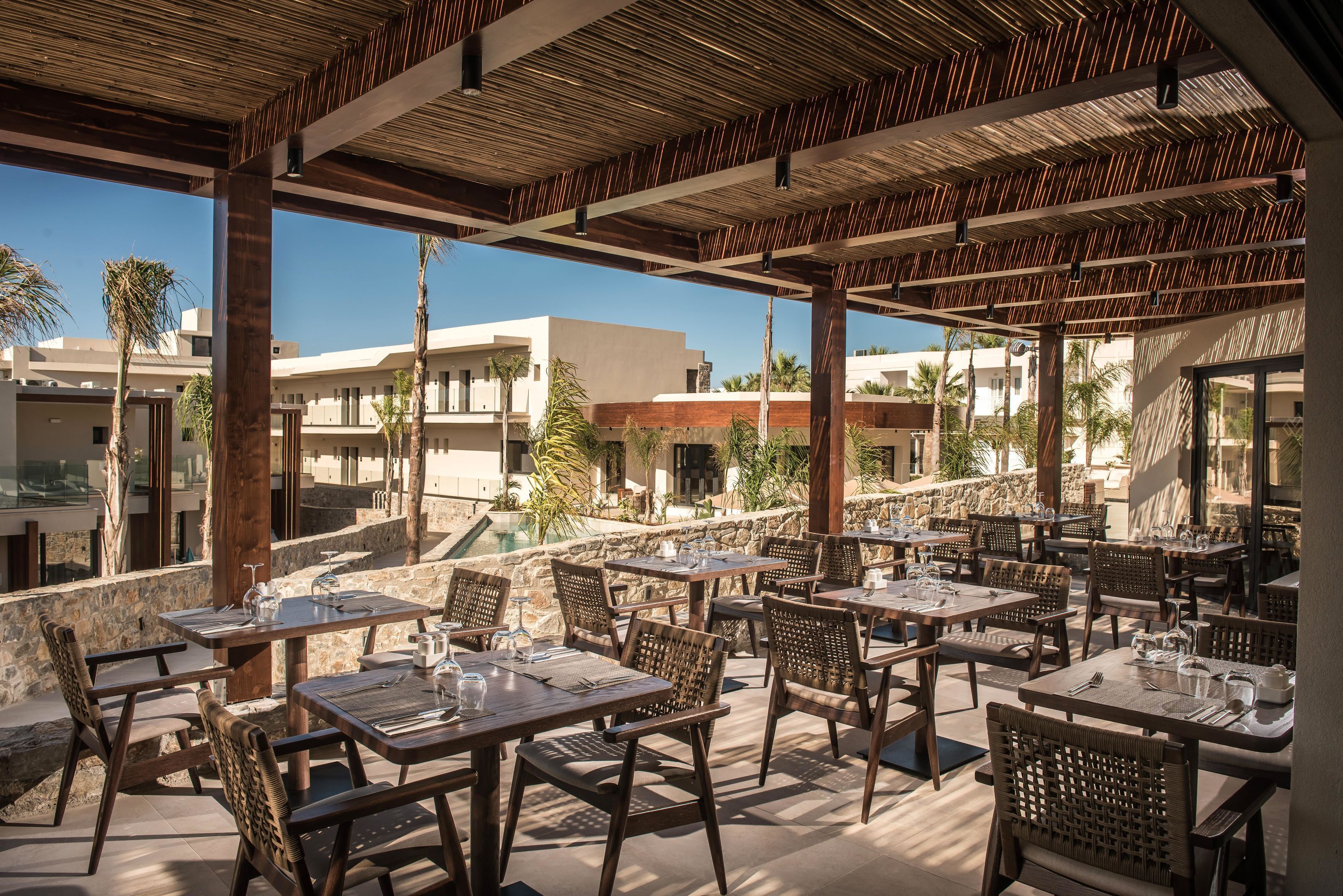 An outdoor dining area with tables and chairs, a stone wall, and buildings in the background.