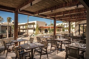 An outdoor dining area with tables and chairs, a stone wall, and buildings in the background. - Noble Hotel Suites - Adults Only (Hersonissos)