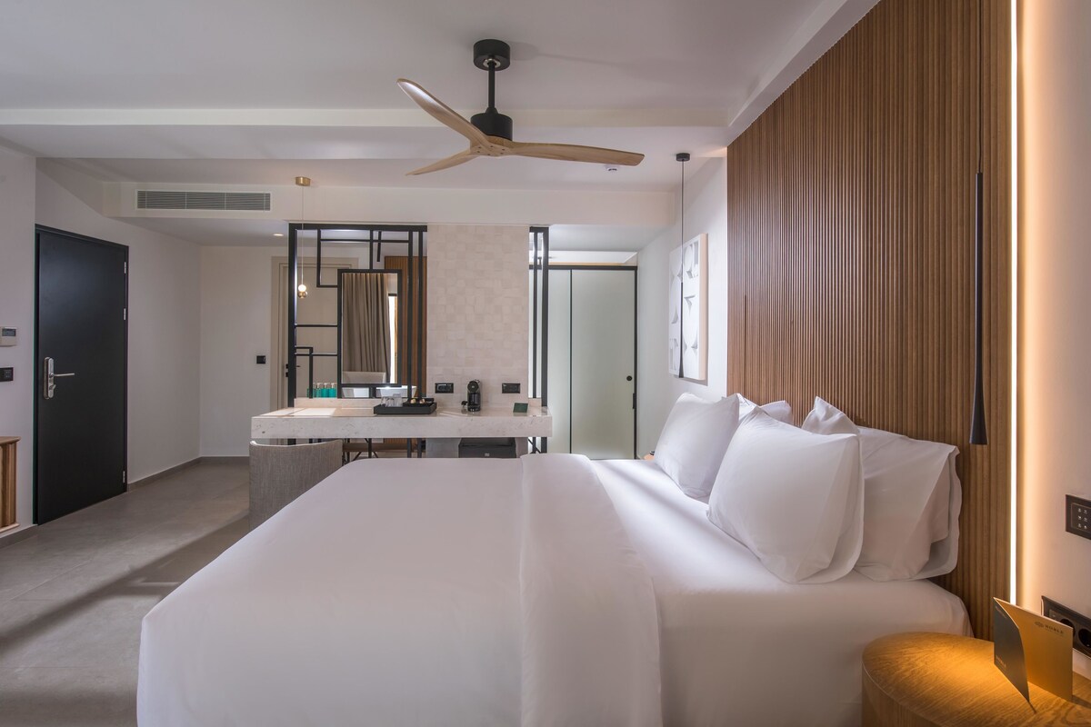 A modern hotel room with a large bed, a desk with a mirror, a ceiling fan, and a wooden panel wall.