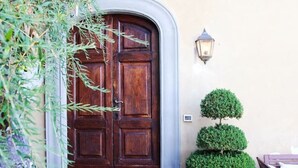 Una puerta doble de madera con diseño tradicional, una planta de boj en maceta a su lado y una lámpara de pared.