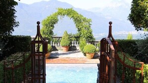 Zona de piscina al aire libre con un portón de madera que da a la piscina, rodeada de plantas en macetas y vistas a montañas.