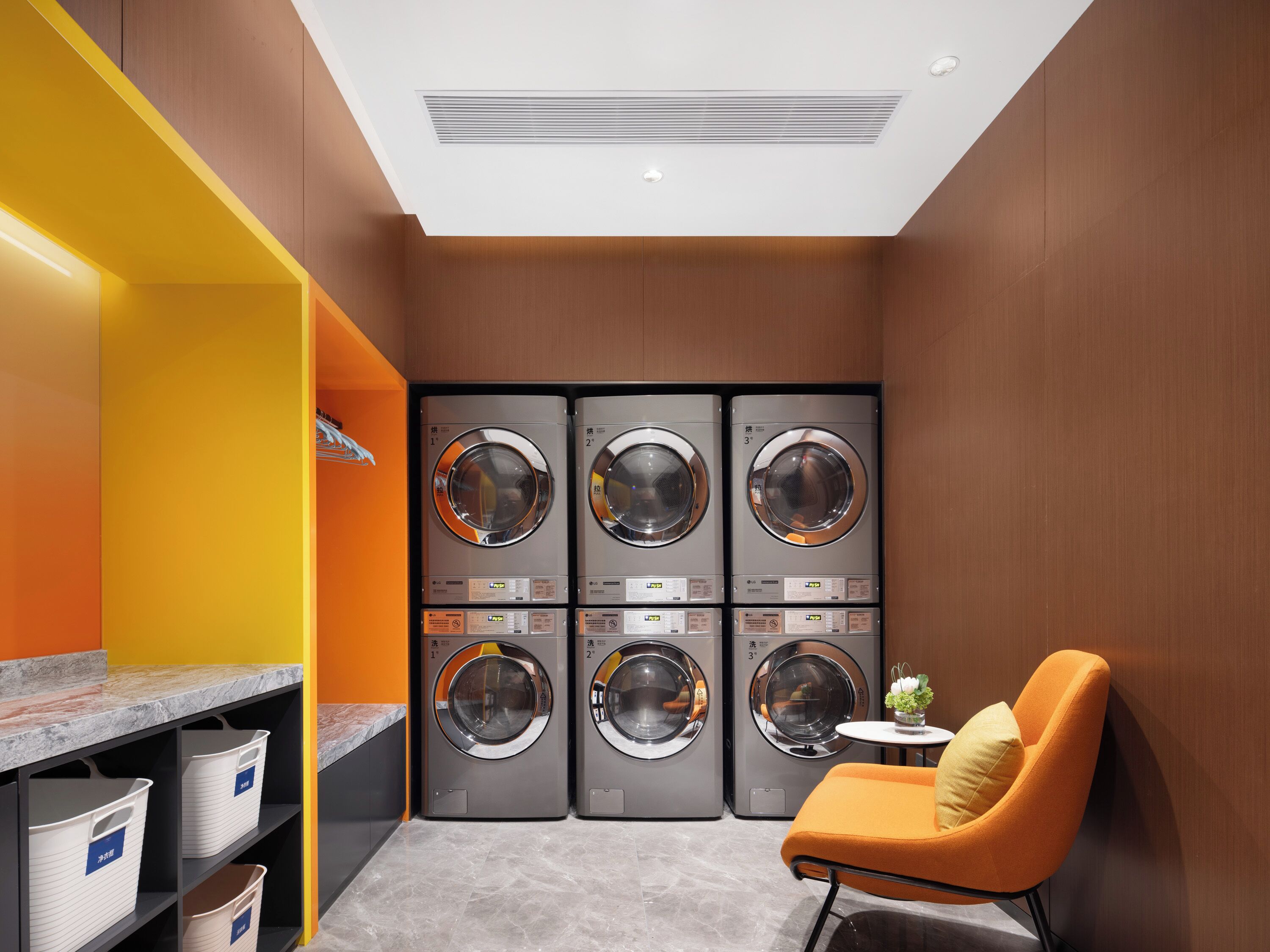 A laundry room with three stacked washing machines, a small table with a vase, and a chair.