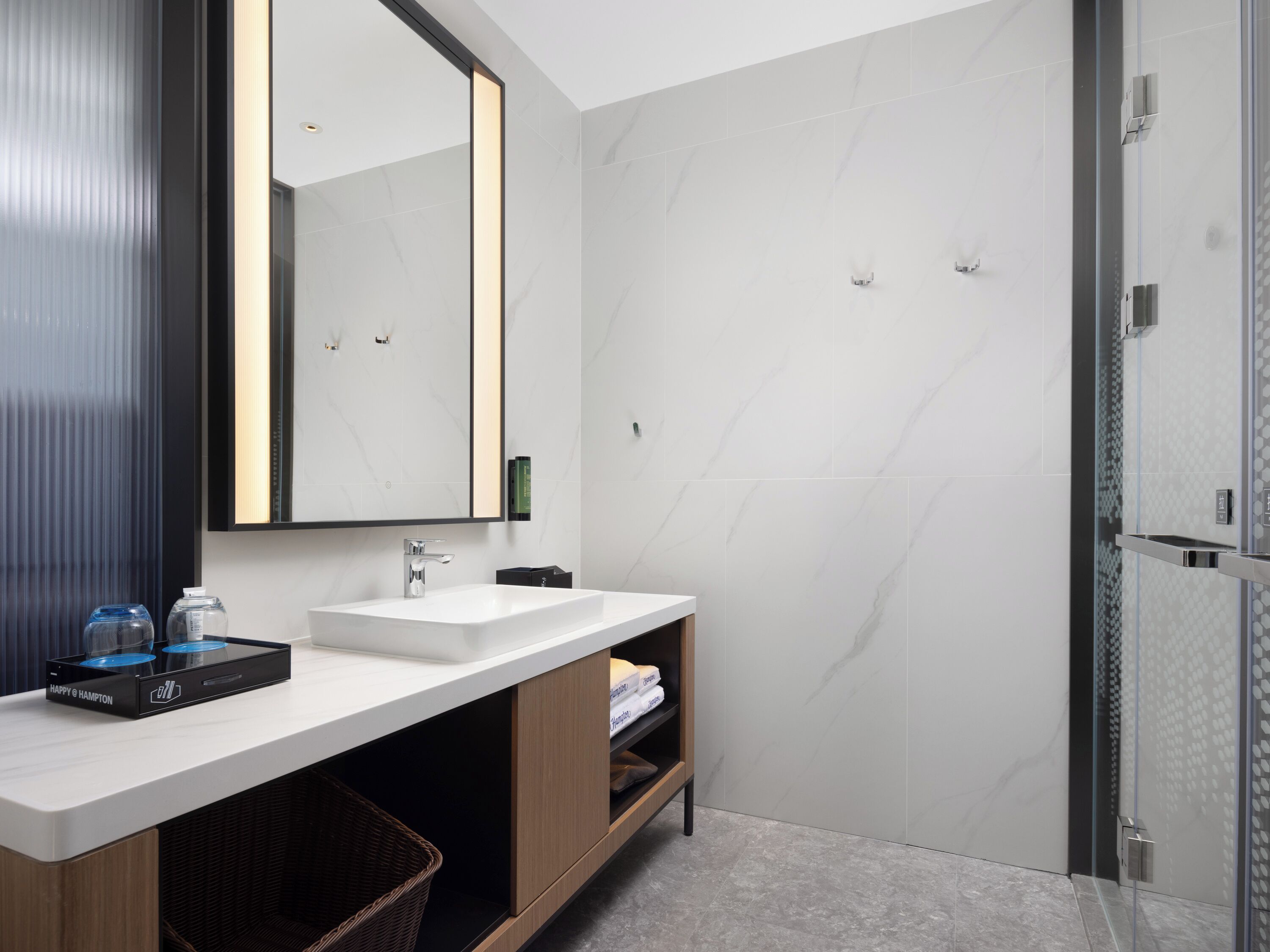 A modern bathroom with a large mirror, white marble walls, a white sink, and a glass shower enclosure.
