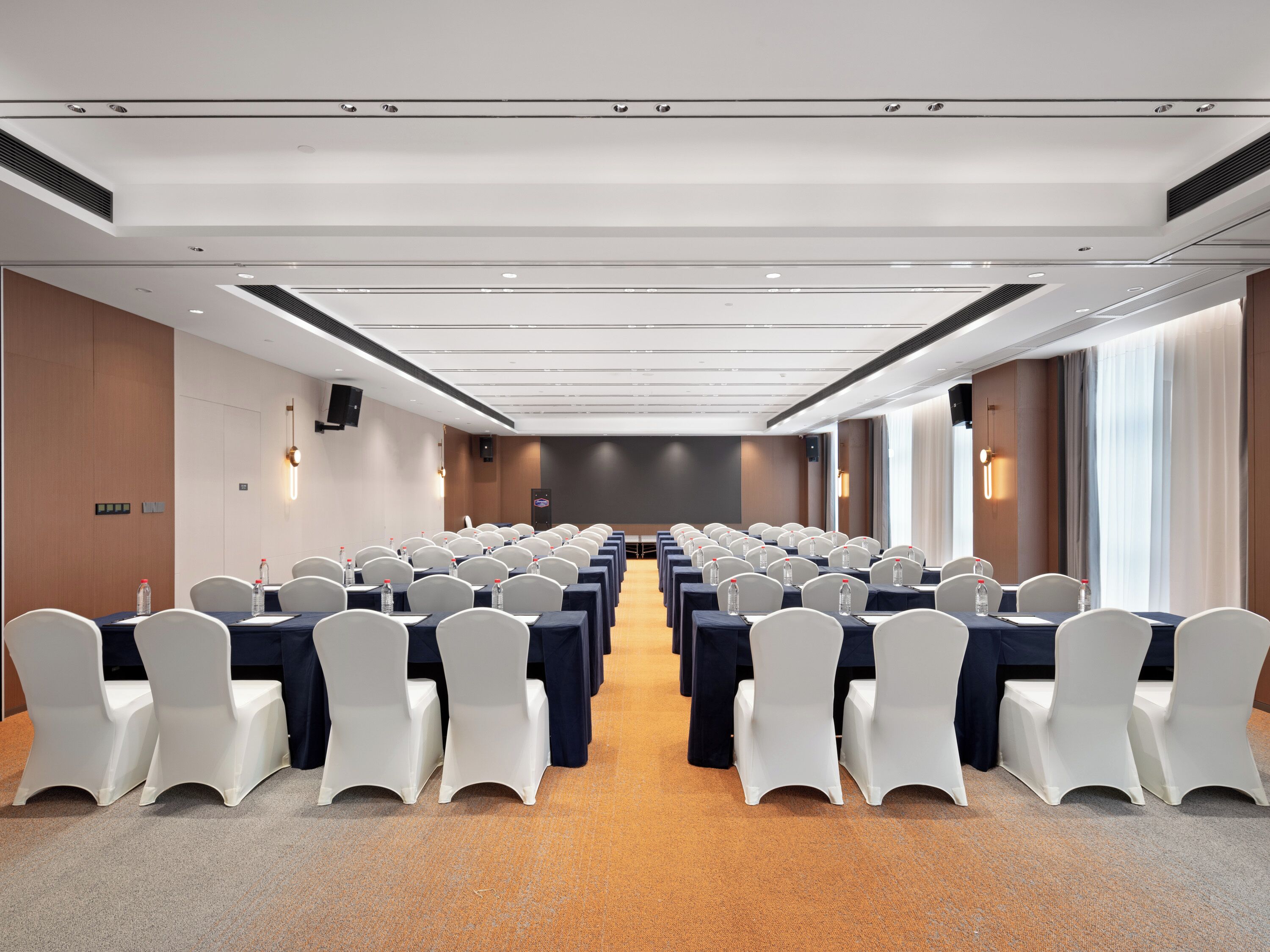 Un salón de conferencias con filas de mesas y sillas, equipado con botellas de agua y un atril.